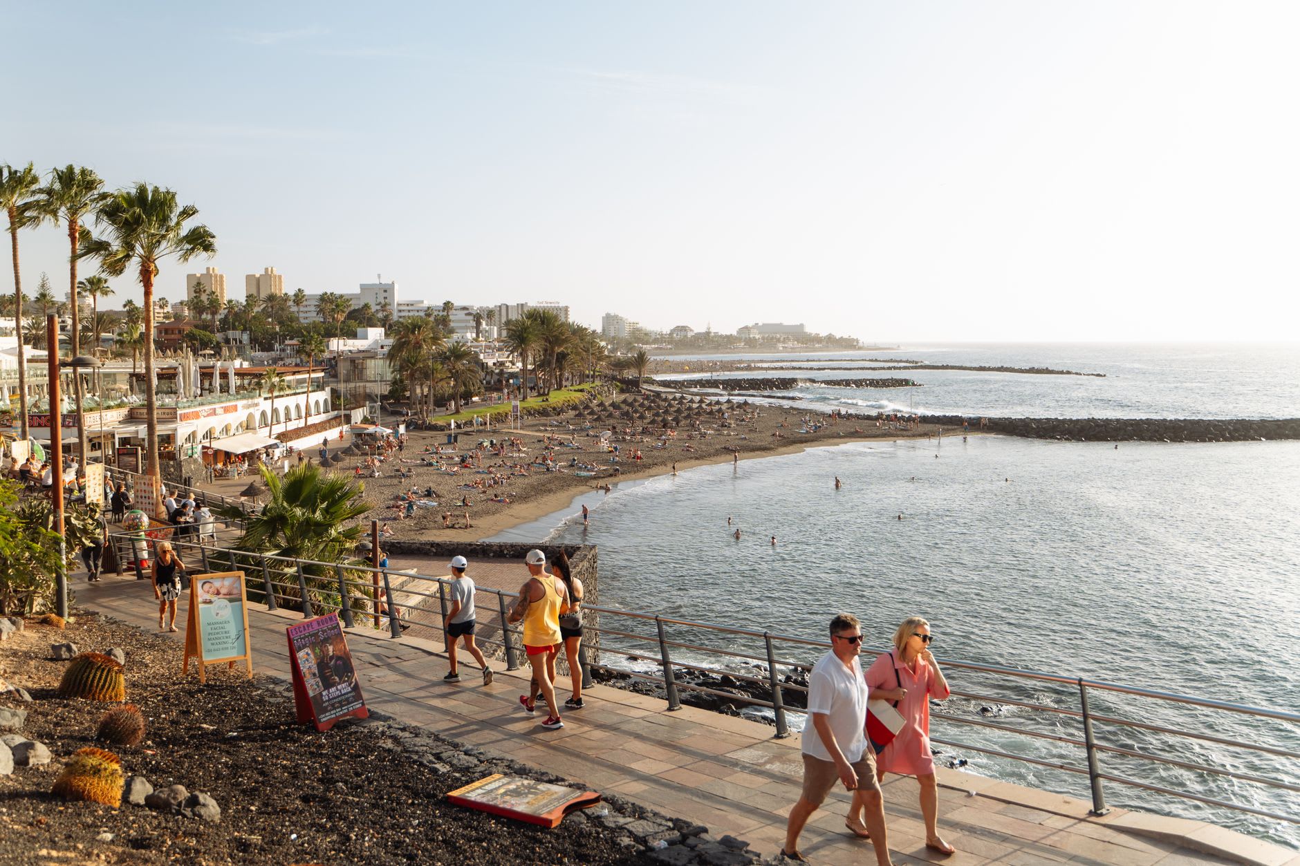 Promenade en bord de mer d'une station balnéaire, plage de sable sombre où quelques personnes se baignent au soleil de fin d'après-midi