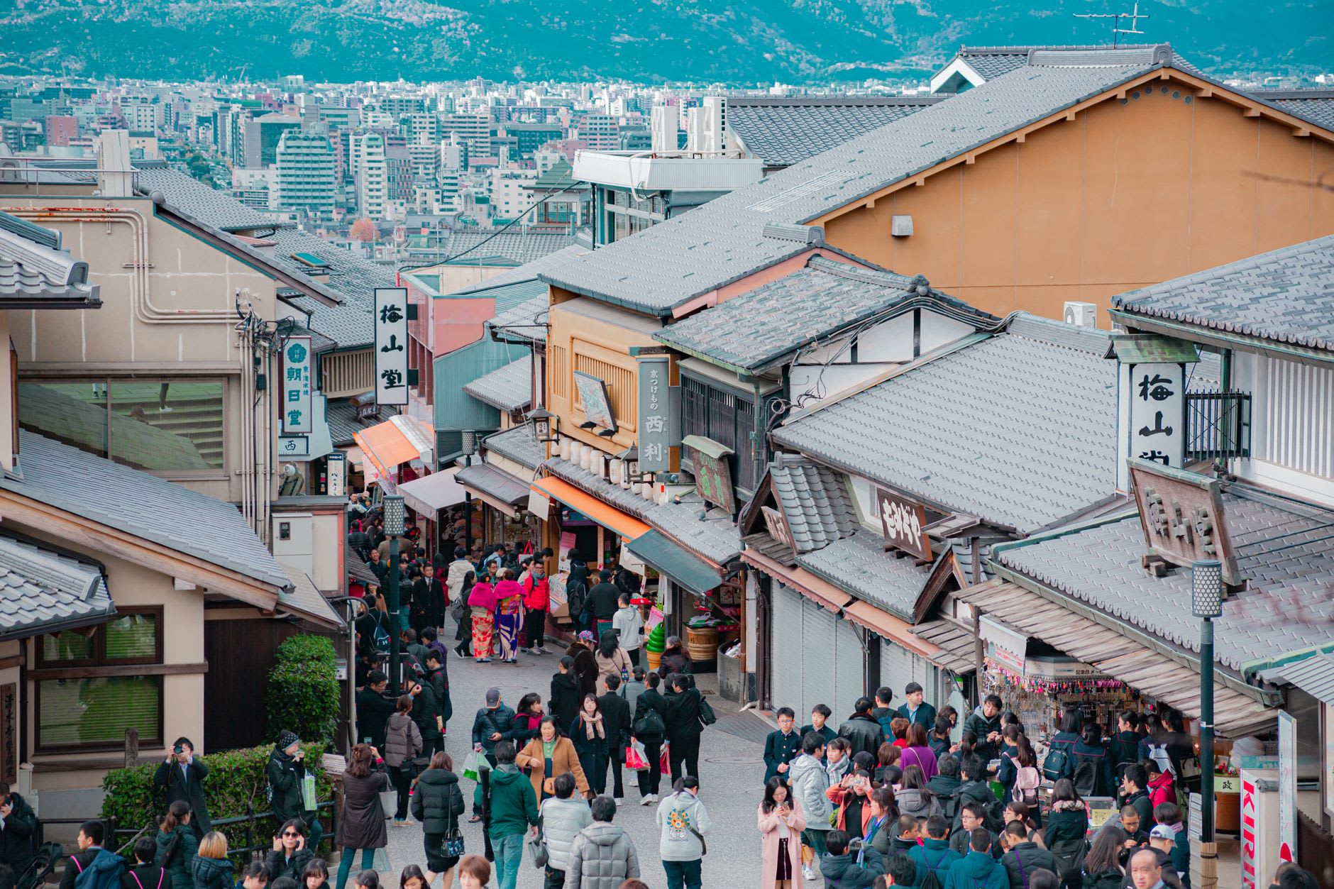 Foule dense de visiteurs remontant une rue commerçante en pente de Kyoto bordée d’échoppes, ville et montagnes au fond sous un ciel clair