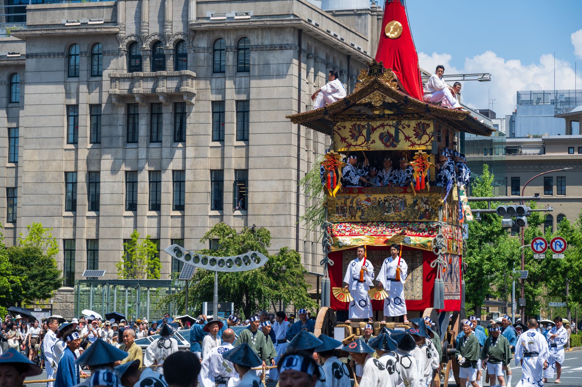 Char de festival japonais haut et doré surmonté d’un mât rouge, porteurs en tuniques bleues et blanches dans une rue de Kyoto sous un ciel d’été