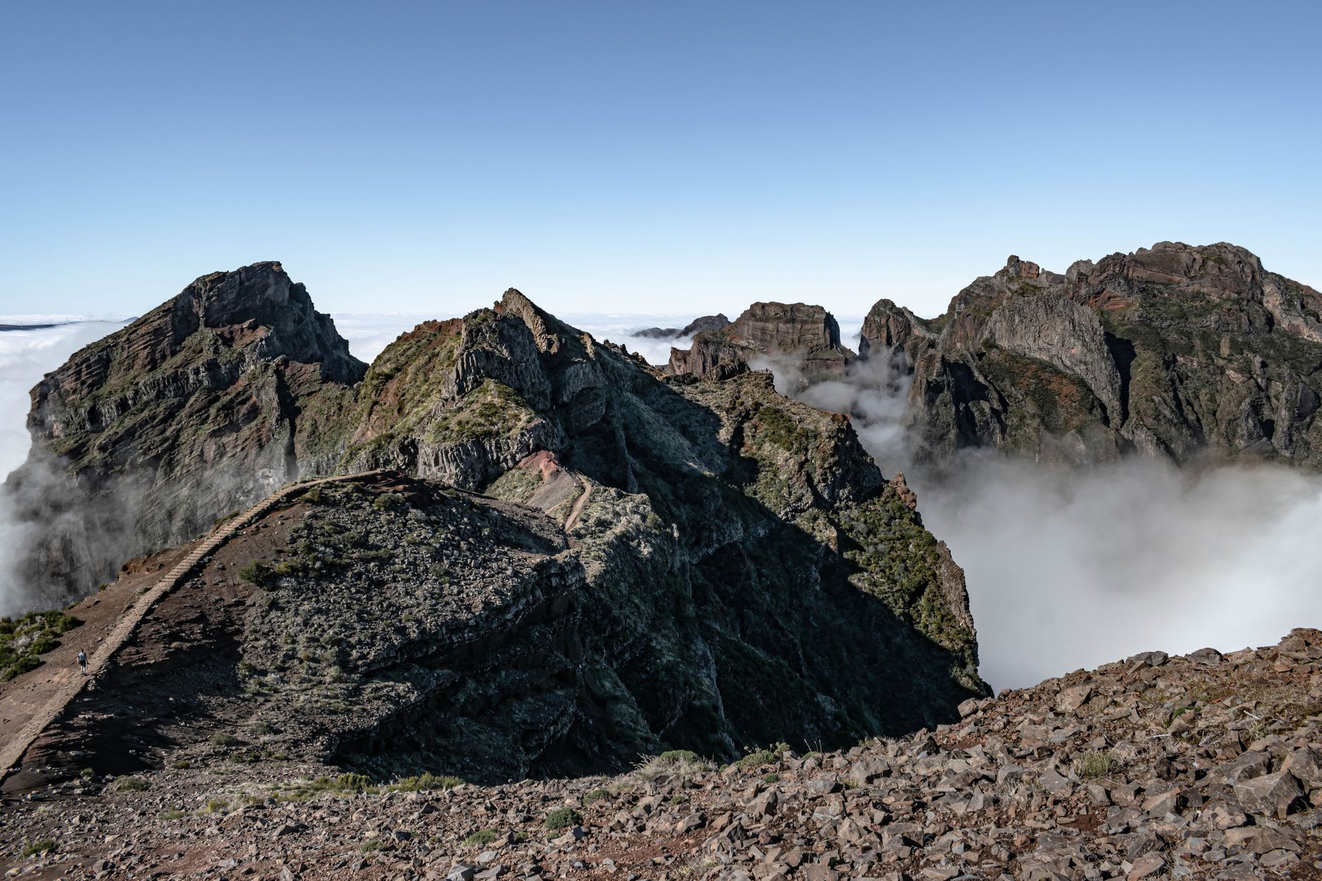 Enfilade de pics volcaniques déchiquetés émergeant d’une mer de nuages sous un ciel bleu, avec un sentier de crête à flanc de montagne