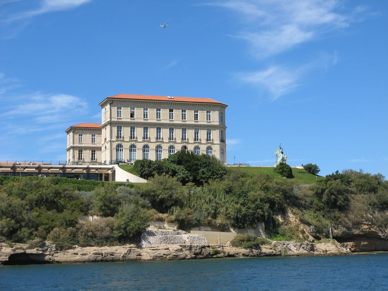 Bâtiment classique à toit de tuiles dominant une côte rocheuse basse, statue de bronze à droite