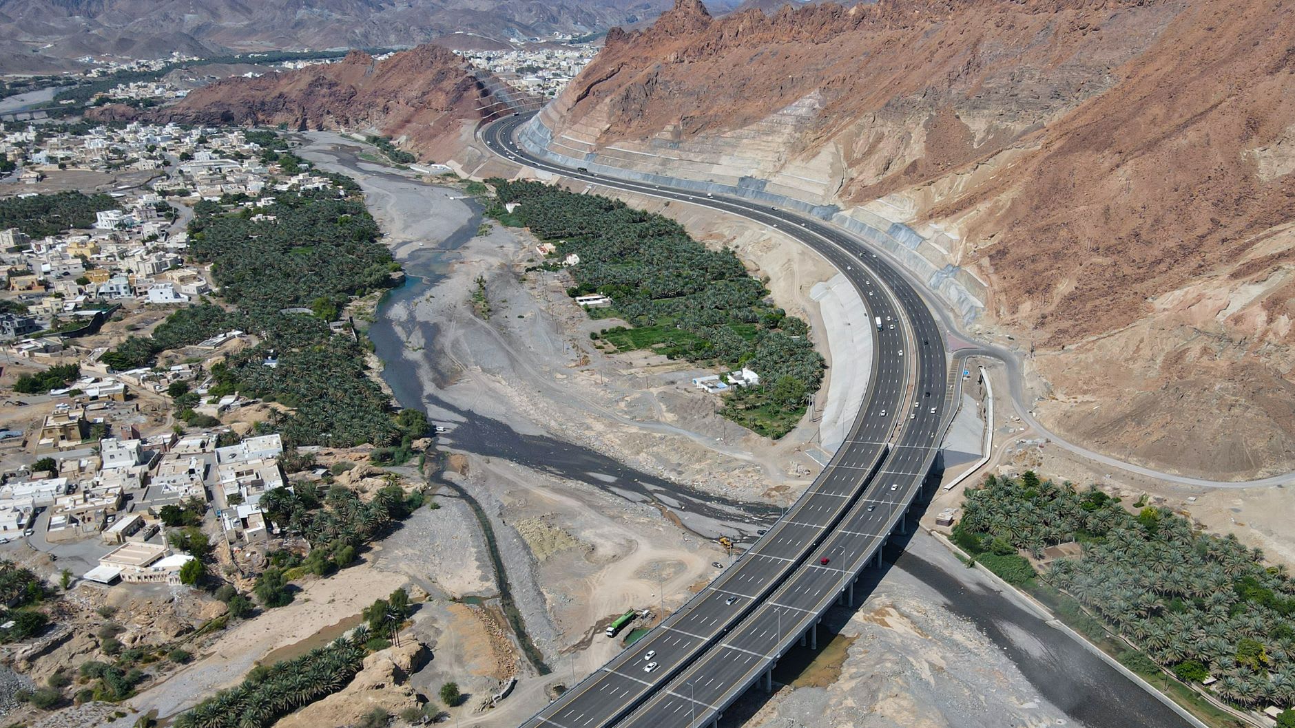 Vue aérienne d’une autoroute franchissant sur pilotis un large lit de rivière au filet d’eau résiduel, bordé de palmeraies