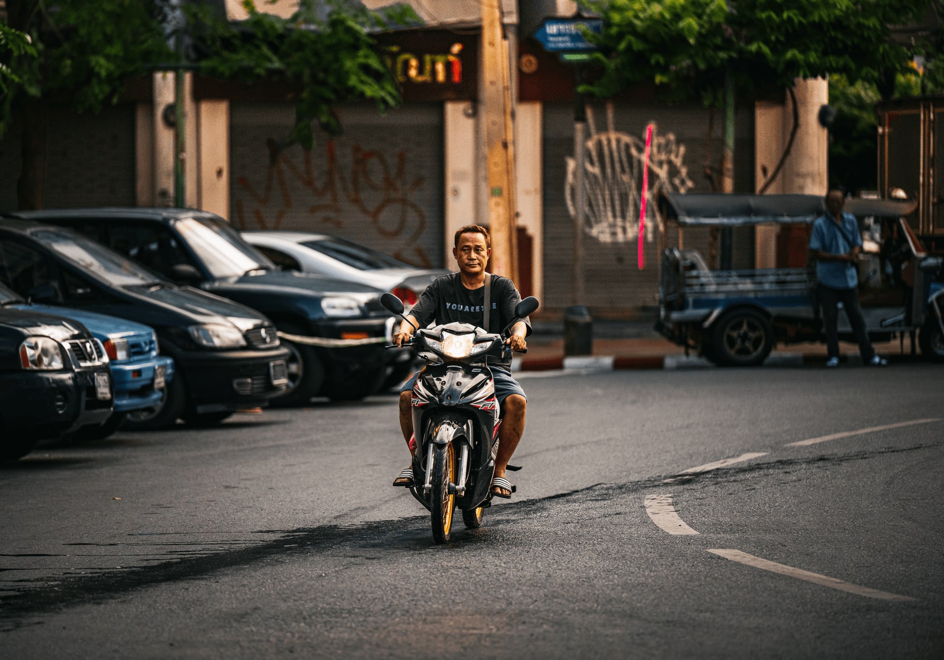 Homme circulant tête nue sur un scooter dans une rue thaïlandaise, tuk-tuk et voitures à l’arrêt en arrière-plan