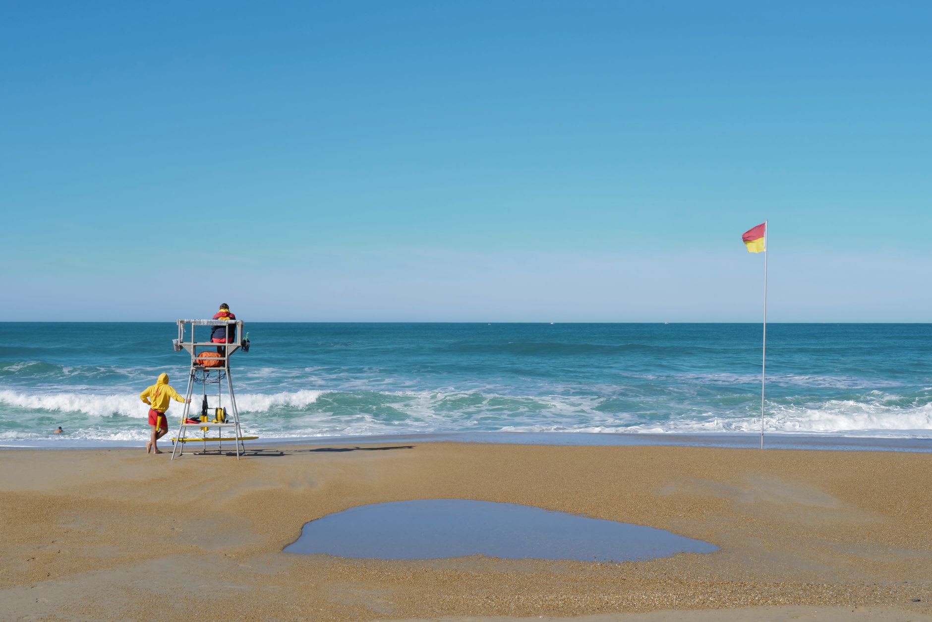 Sauveteur assis en vigie près d’un drapeau rouge et jaune planté au bord de l’eau, face aux vagues