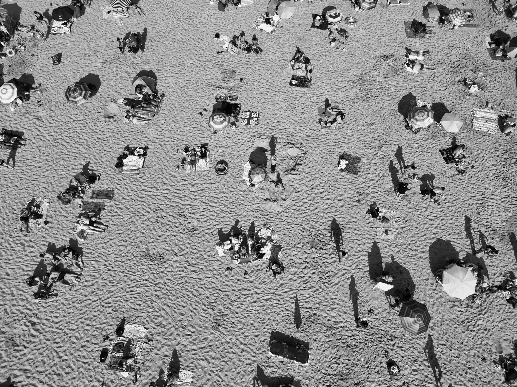 Plage de sable vue du ciel, saturée de parasols et de baigneurs aux ombres longues, en noir et blanc