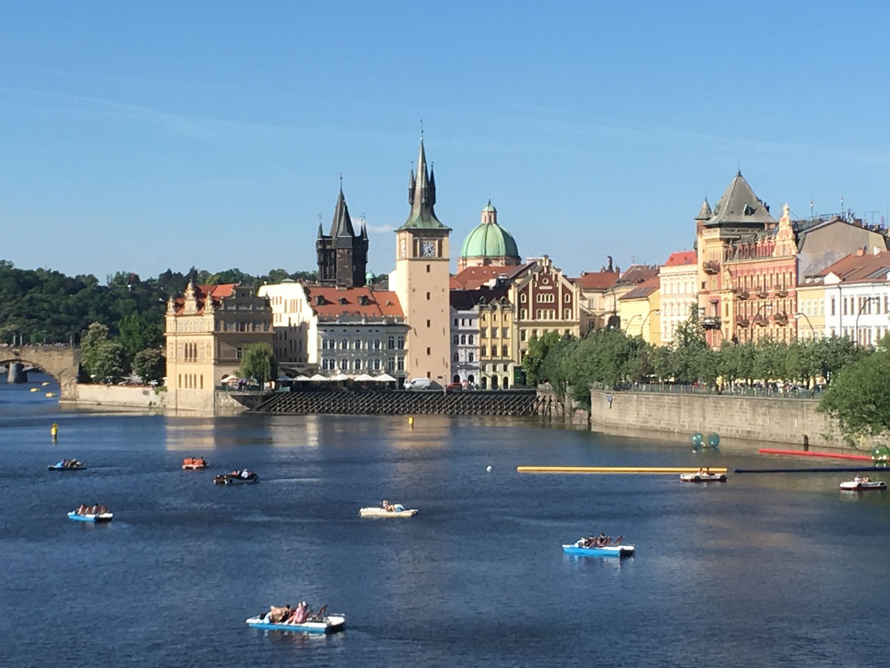 Nombreux pédalos bleus chargés de passagers sur la Vltava ensoleillée devant une rangée d’immeubles anciens et une coupole verte