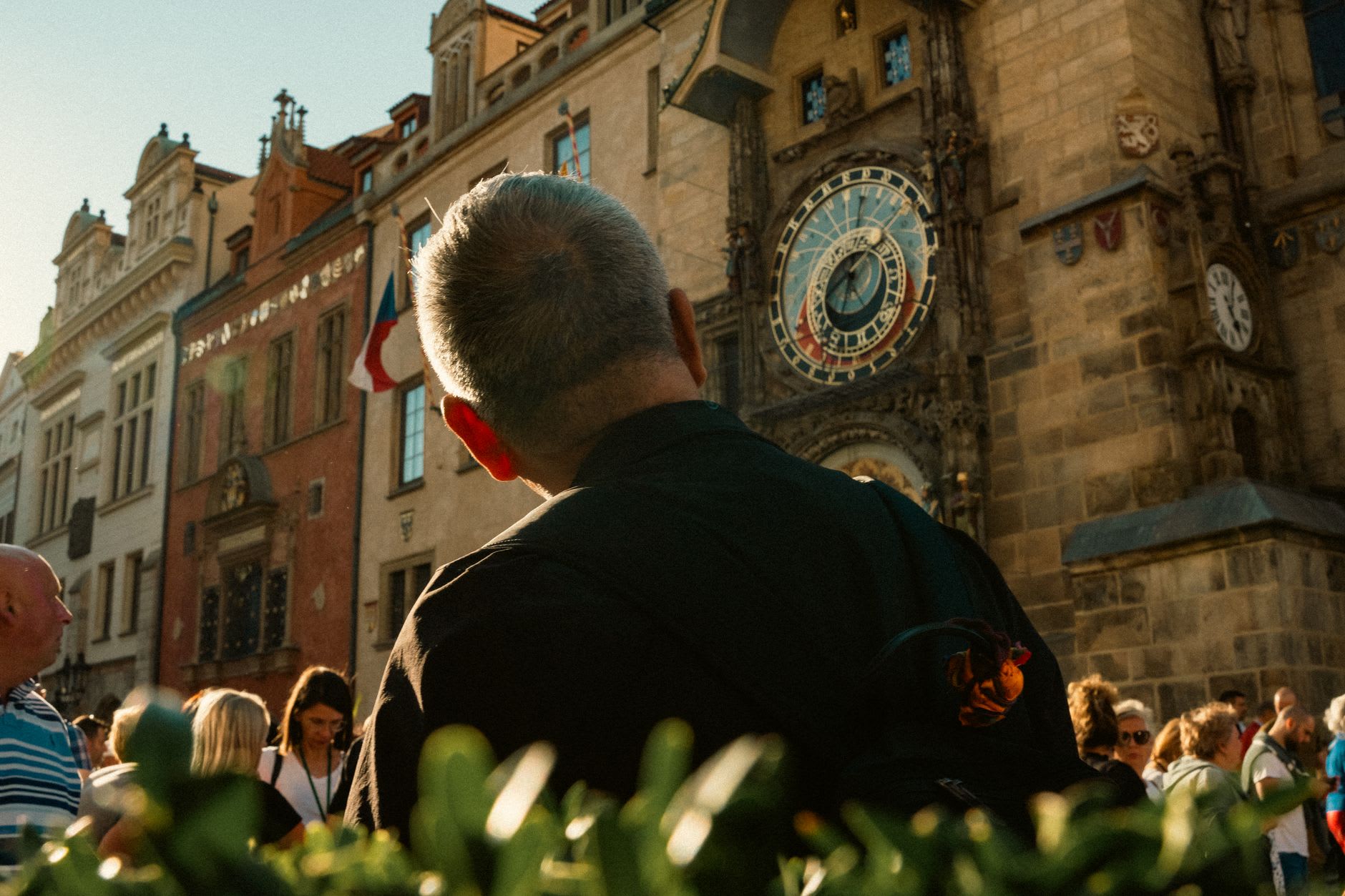 Homme de dos observant l’horloge astronomique médiévale au milieu d’une foule de visiteurs, façade de pierre dorée par la lumière du soir