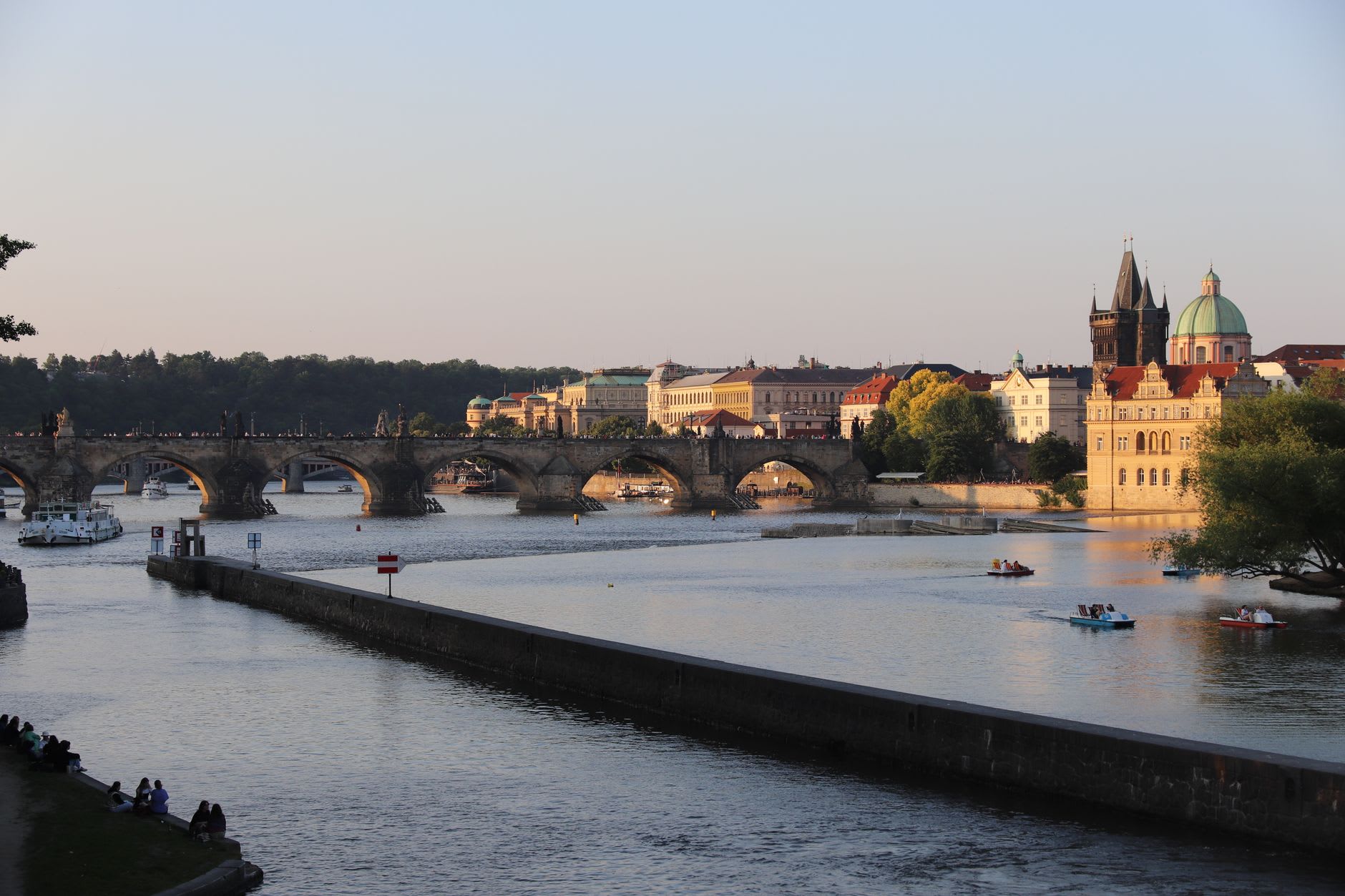 Pont Charles doré par la lumière du soir au-dessus de la Vltava, pédalos sur l’eau et promeneurs assis sur le quai