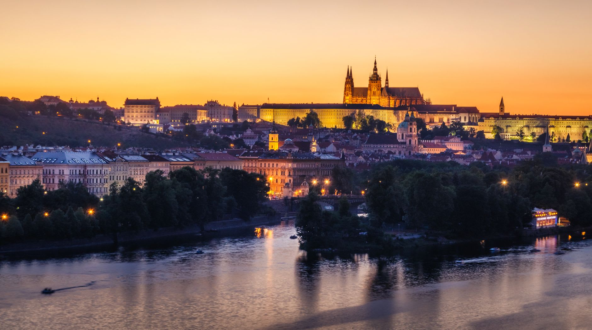 Château de Prague et cathédrale Saint-Guy illuminés au coucher du soleil, ciel orange et reflets sur la Vltava en contrebas