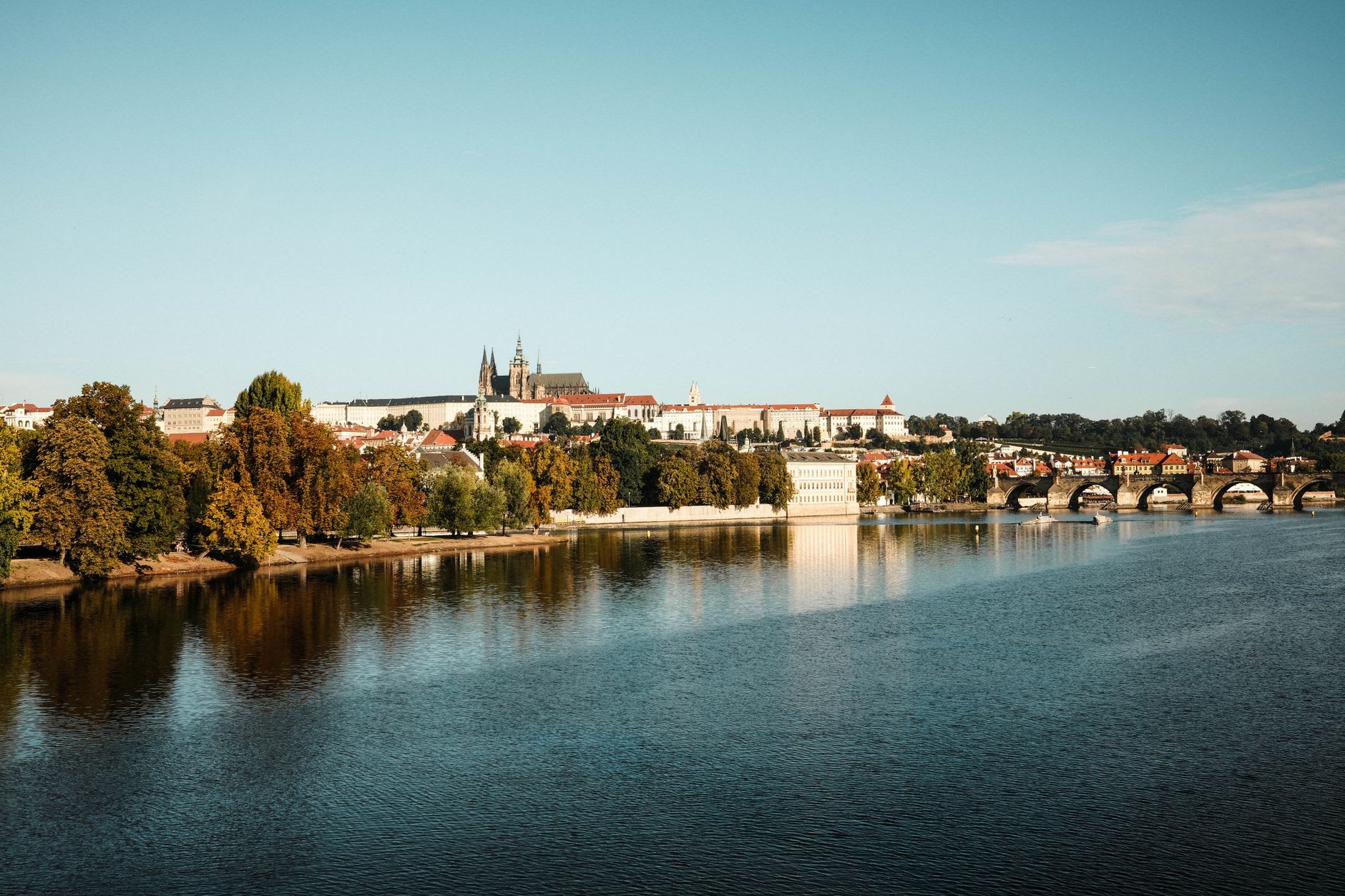 Les arches de pierre du pont Charles et le château au-dessus de la Vltava, des arbres roux sur la rive et leurs reflets dans l’eau calme