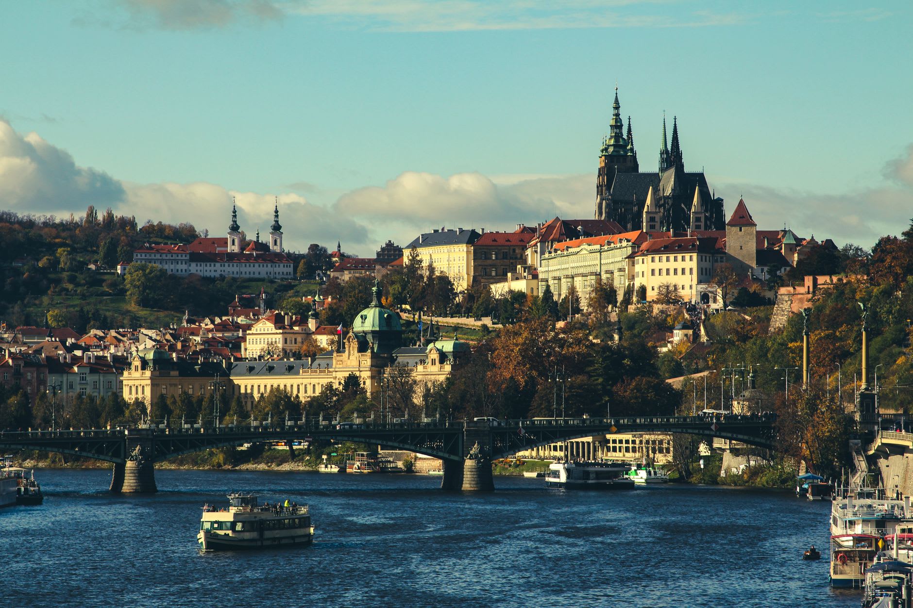 Le château de Prague et sa cathédrale gothique au-dessus de la Vltava, un pont et des bateaux de promenade au premier plan dans une lumière chaude de fin d’après-midi