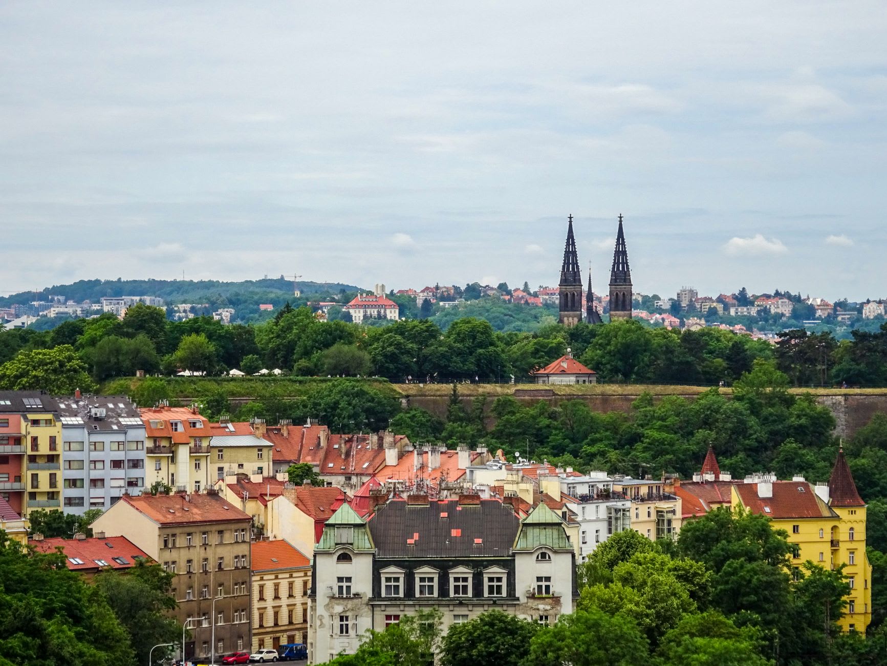 Toits de tuiles rouges d’un quartier de Prague dominés au loin par les deux flèches noires de la basilique de Vyšehrad sur sa colline boisée