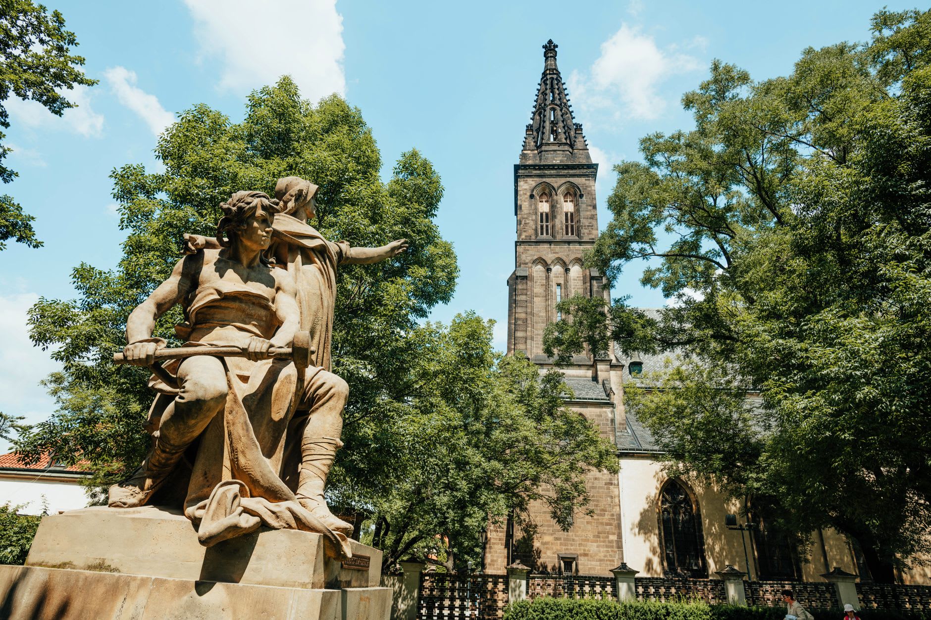 Statue de pierre dans un parc arboré devant la haute tour néogothique de la basilique de Vyšehrad, sous un ciel d’été bleu