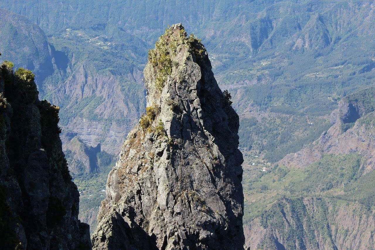 Piton rocheux acéré dominant un cirque encaissé, petites cases dispersées sur les planèzes vertes tout au fond de la vallée