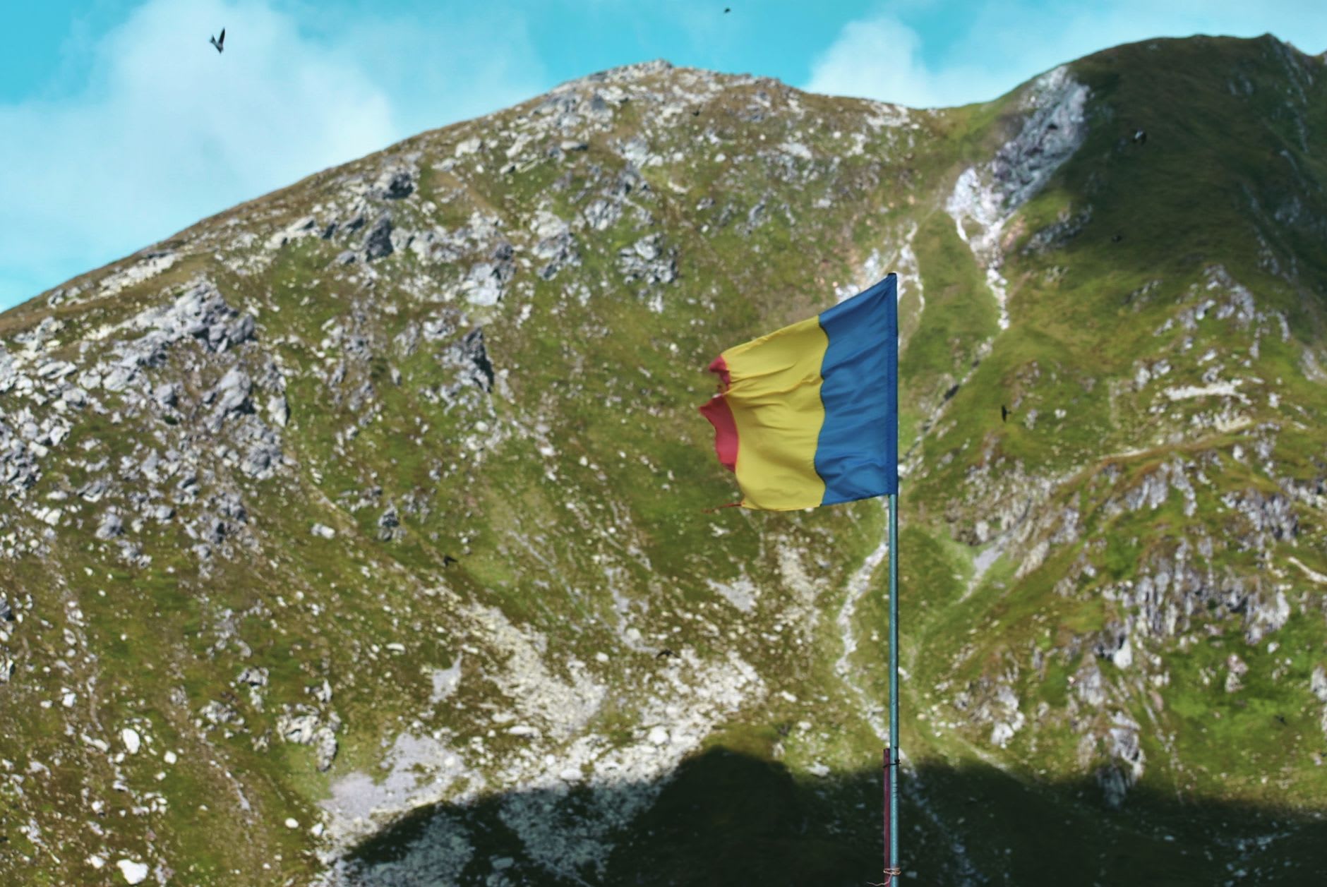 Drapeau roumain bleu, jaune et rouge planté sur un mât devant un versant rocheux des Carpates, le tissu usé par le vent