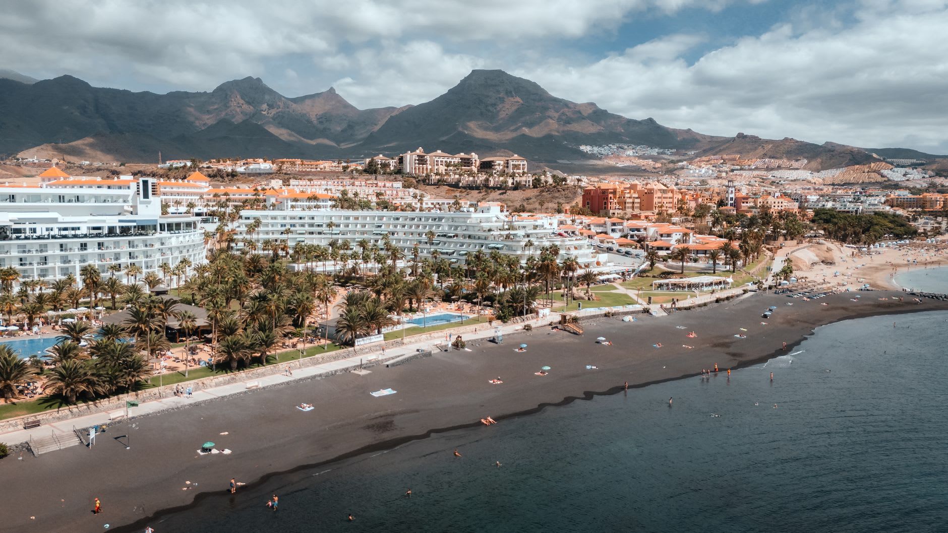 Vue aérienne d’une plage de sable noir bordée de grands hôtels et de palmiers, baigneurs épars et montagnes à l’arrière sous un ciel nuageux