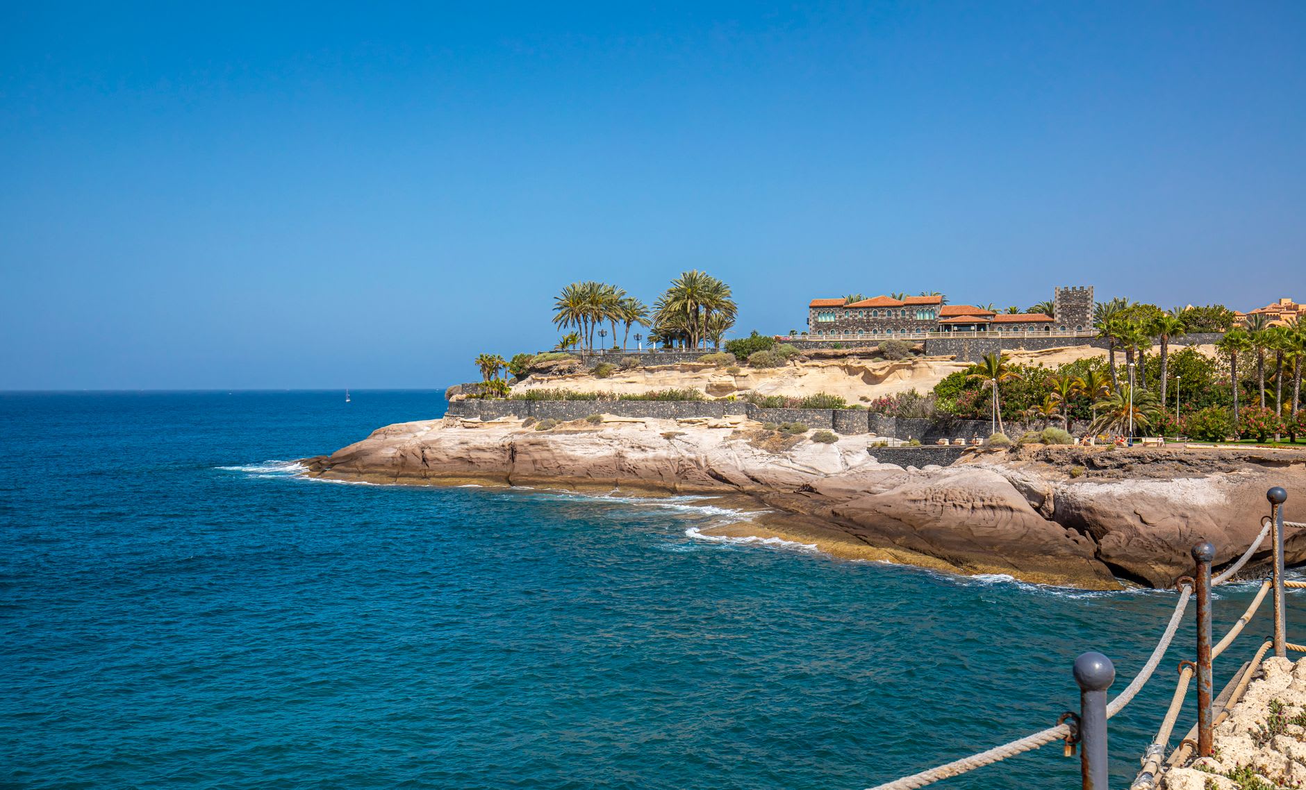 Pointe rocheuse ocre bordée de palmiers et d’un hôtel, léchée par une mer bleu profond sous un ciel dégagé, sur la côte sud de l’île