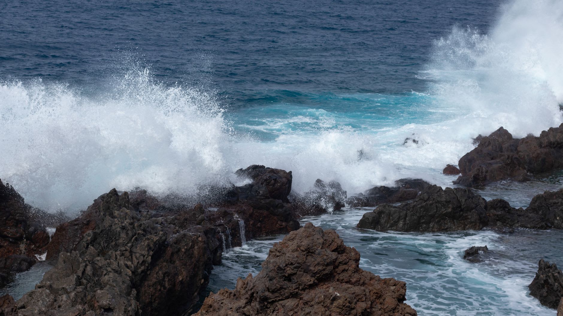 Vagues explosant en gerbes d’écume blanche contre des rochers volcaniques déchiquetés, l’océan bleu sombre à l’arrière-plan