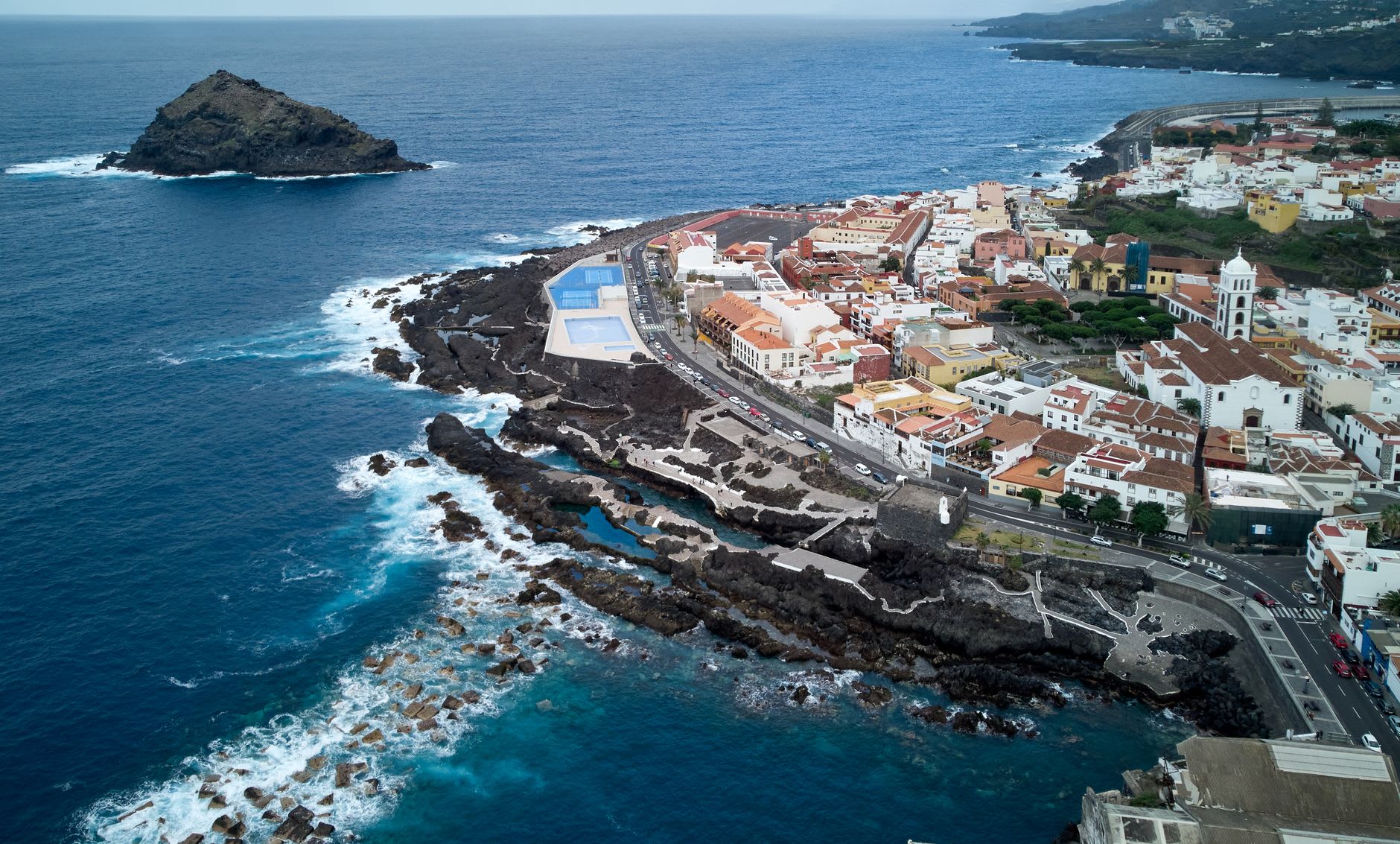 Vue aérienne d’une ville blanche posée sur une côte de lave noire, un rocher-îlot au large et des bassins d’eau de mer creusés dans la roche