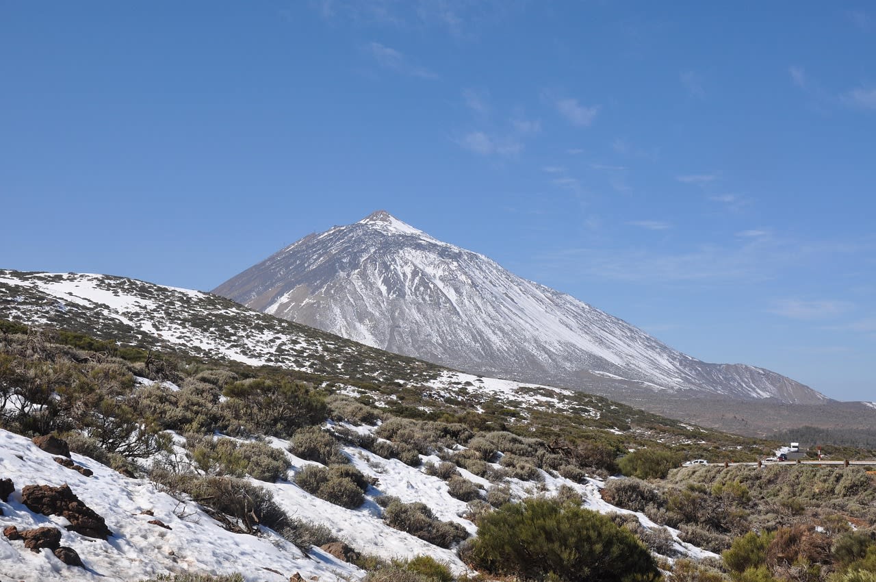 Sommet enneigé du Teide dominant un plateau de lave couvert de plaques de neige, avec une route de montagne et des véhicules sur la droite