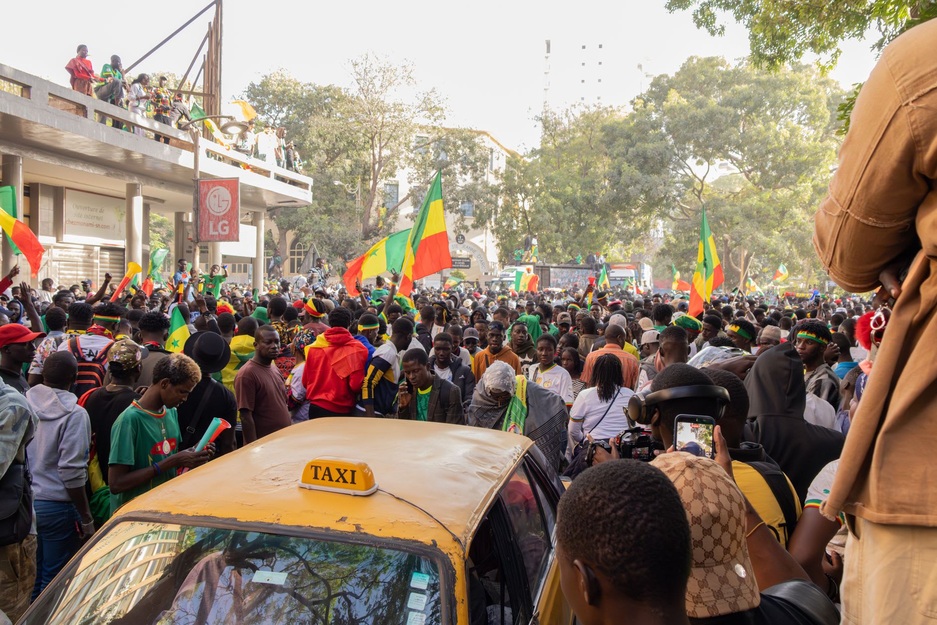 Foule dense agitant des drapeaux sénégalais autour d’un taxi jaune dans une avenue