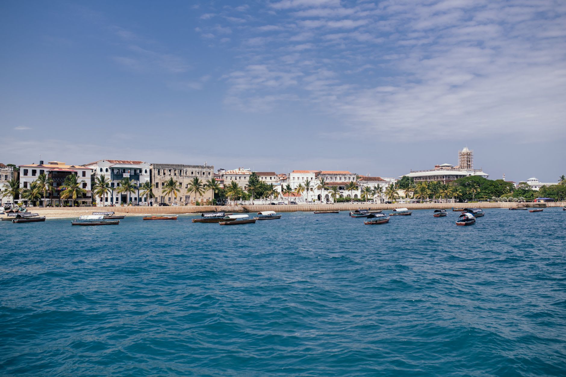 Front de mer de Stone Town vu depuis l’eau, boutres amarrés devant des demeures blanches sous un ciel voilé de chaleur