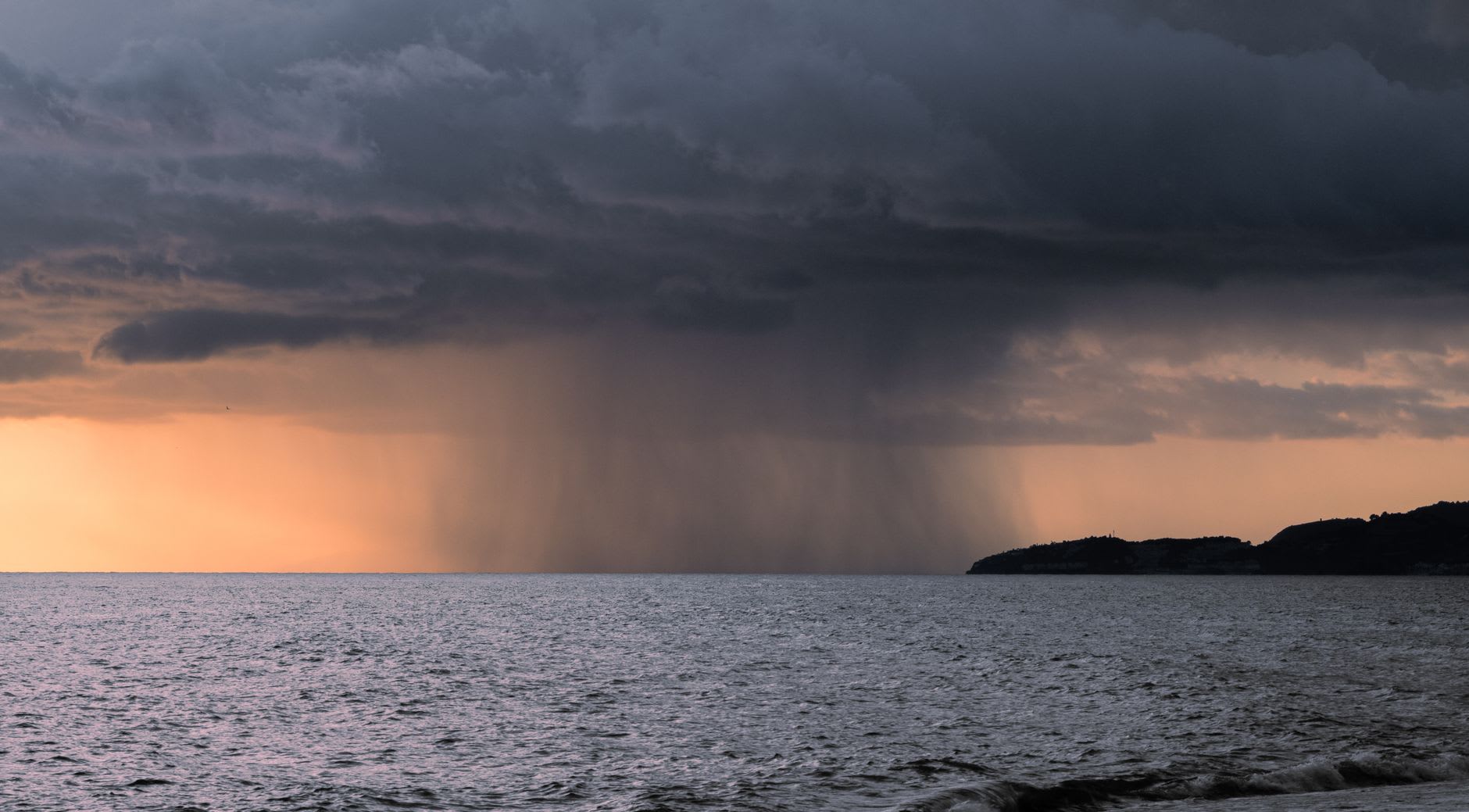 Une averse tropicale tombe en rideau serré depuis un nuage sombre sur la mer, lumière dorée du couchant à gauche
