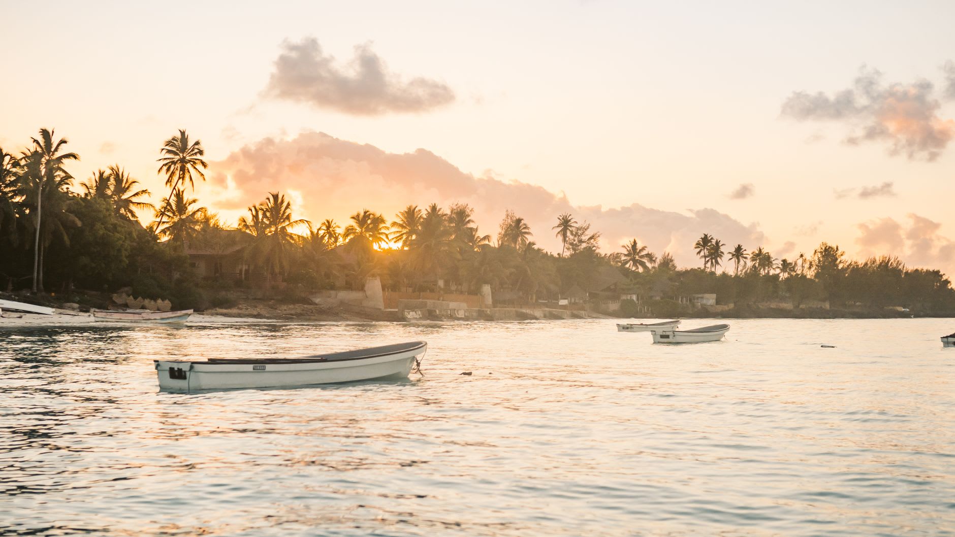 Coucher de soleil orange sur une baie calme, plusieurs barques de pêche au mouillage devant une côte plantée de cocotiers