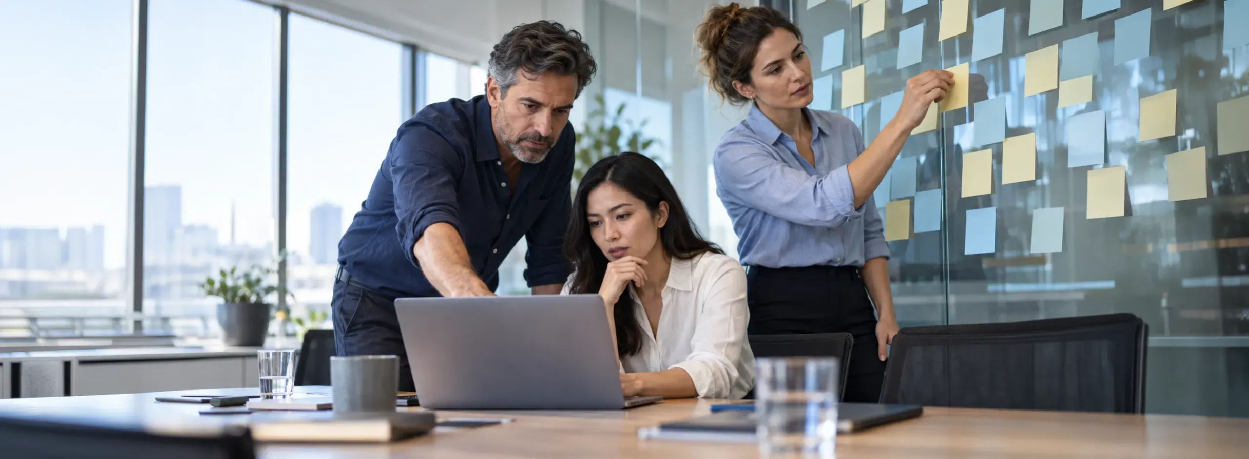 Three colleagues reviewing a laptop and arranging sticky notes on a glass planning board