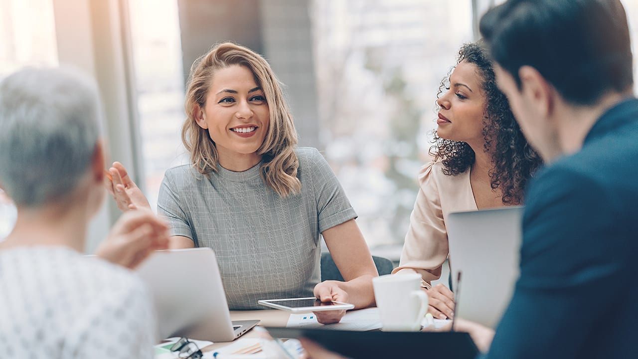 woman working on laptop and holding tablet smiles during meeting with 3 colleagues