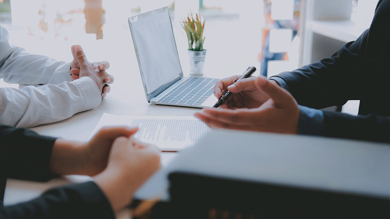 gesturing man holds a pen over documents facing 2 other people, as if they are signing an agreement