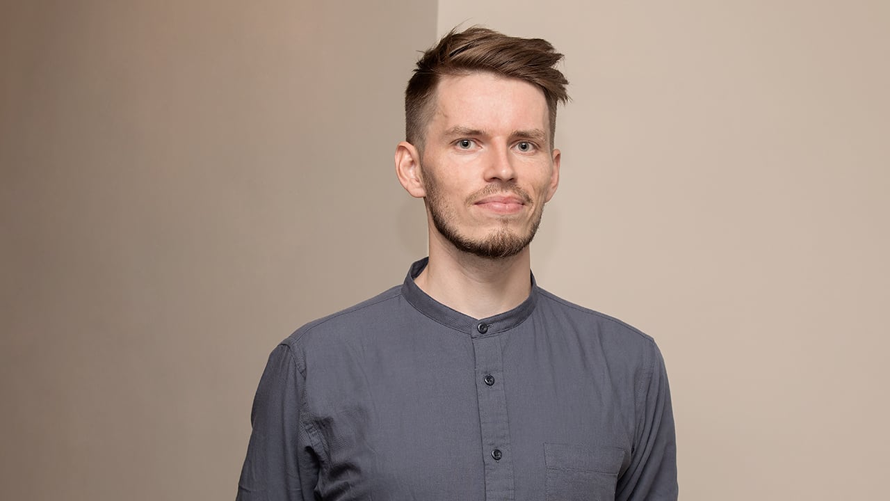 Young man in grey shirt, with a styled haircut and beard, smiles at camera