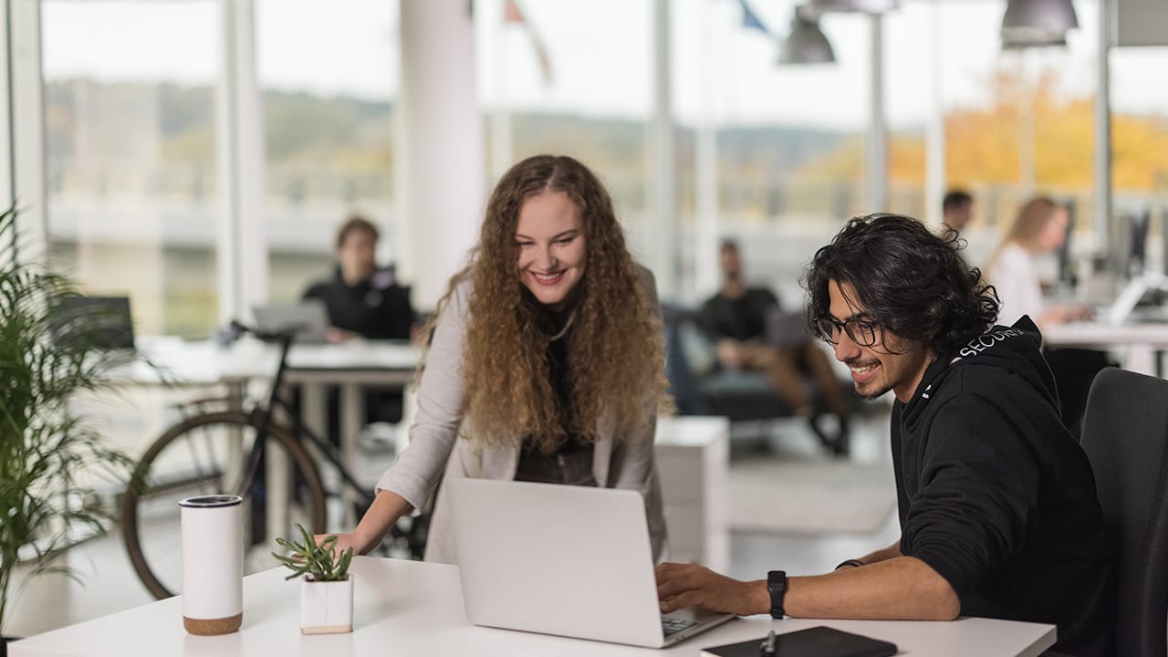 man in nord hoodie and woman in a a busy office look at laptop and smile