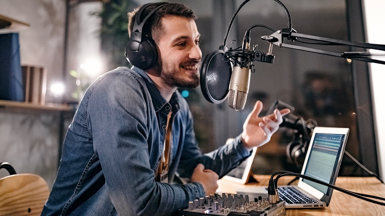 headphone wearing podcast host in denim shirt smiles and gestures while speaking into mic