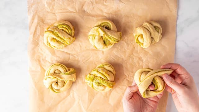 Strekk deigstrimlene litt ut og surr dem rundt hånda slik at du får pistasjknuter. Legg enden under bollen. Ha bollene på et bakebrett med bakepapir.
