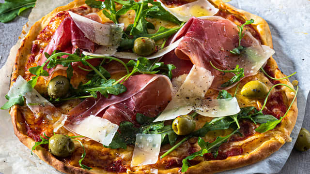 Fordel parmaskinke, høvlet parmesan, oliven og rucculasalat over den varme bunnen og server.