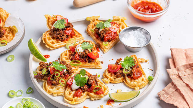 Finhakk vårløk og del cherrytomatene i to. Topp vaflene med tacorestene, vårløk, cherrytomater og revet ost.