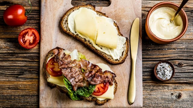 Smør aioli på begge skivene. Legg tre skiver ost på den ene skiven og topp med tomatskiver, salat og sprøstekt bacon. Krydre med salt og pepper.