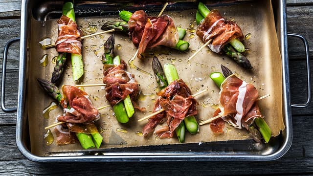 Del aspargesen i to, legg de to delene inntil hverandre og rull litt skinke rundt. Fest skinken med en cocktailpinne og legg aspargesen over i en ildfast form eller på et stekebrett med bakepapir.