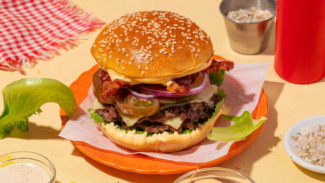 Anrett burgeren lagvis: Ha burgerdressing på bunnen. Topp med romeosalat, to smashburgere, tomat, løk og bacon. Smør litt dressing på toppen av burgerbrødet og legg det på.