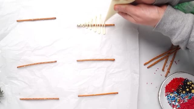 Legg saltstengene på et bakeark med god avstand. Sprøyt en sammenhengende strek med hvit sjokolade nedover saltstangen. Streken skal gå lengre ut på hver side slik at det ser ut som et juletre.
