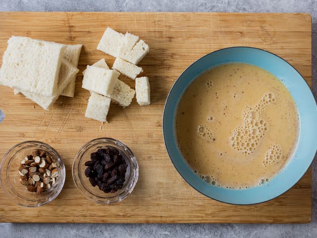 Skjær skorpene av skivene og del dem opp i biter. Pisk sammen egg, melk, sukker og kanel. Tilsett frøene fra vaniljestangen og mesteparten av mandlene og rosinene.