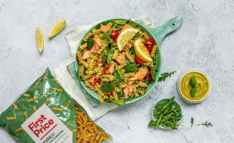 Stor bolle med pastasalat og laks står på et bord. Ved siden av ligger en stor pose med fusilli pasta fra First Price.