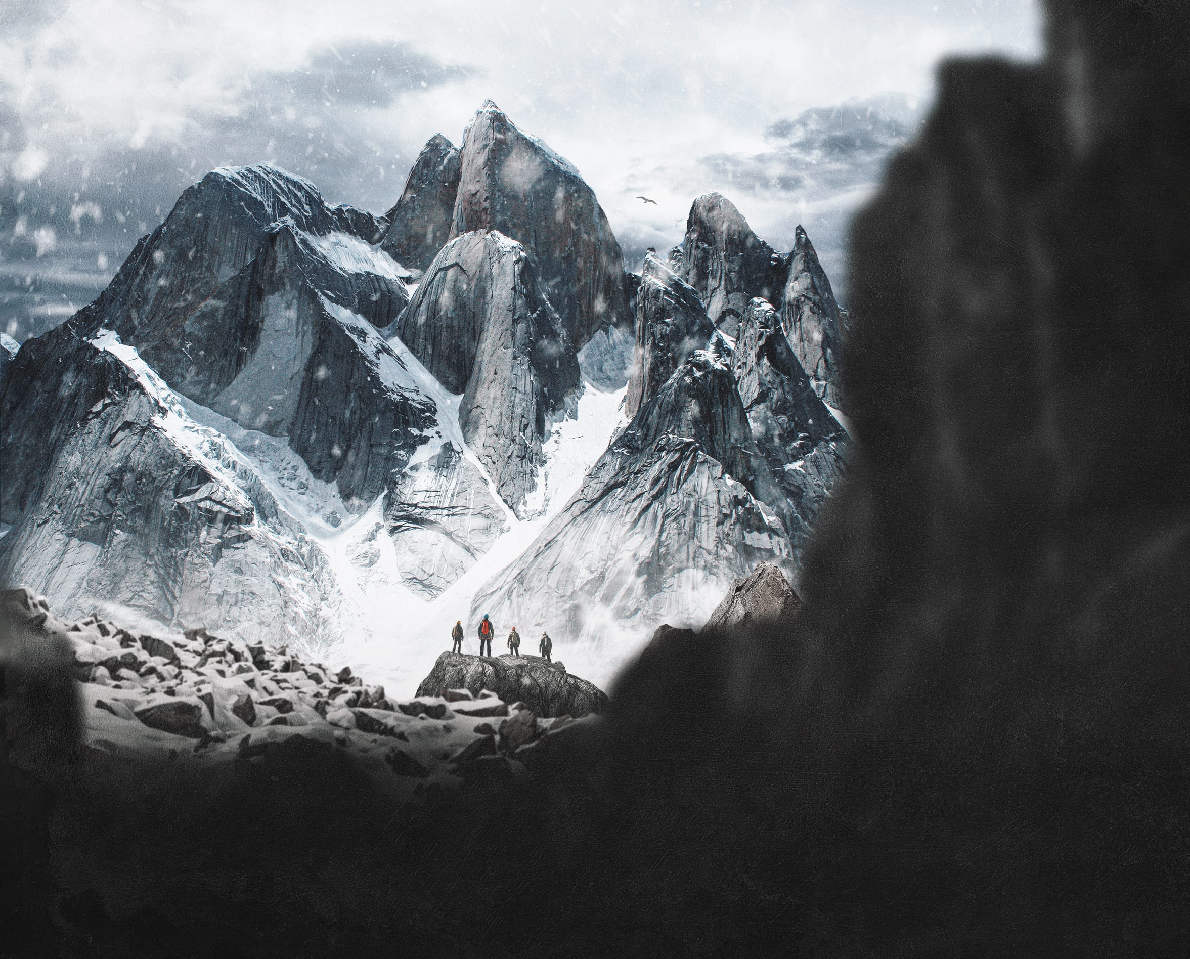 Four hikers in Norrøna apparel stand on a rocky peak with a snow-covered mountain range behind them. Snow is falling.