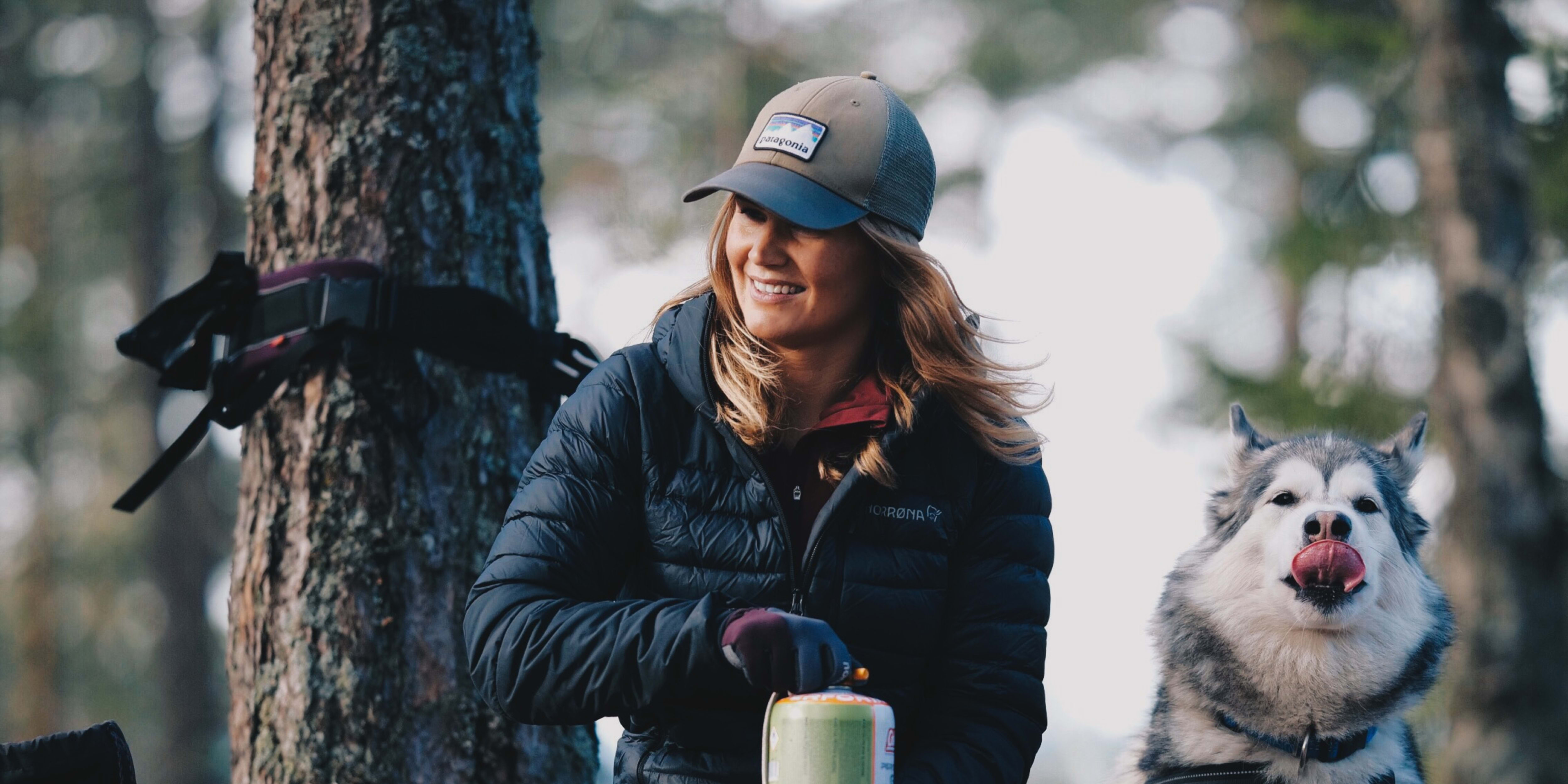 A woman in a black jacket and red pants is crouching in a forest with her dog, a husky. She is holding a green can. The dog is sitting with its tongue out. The background is out of focus, and there are trees in the background.