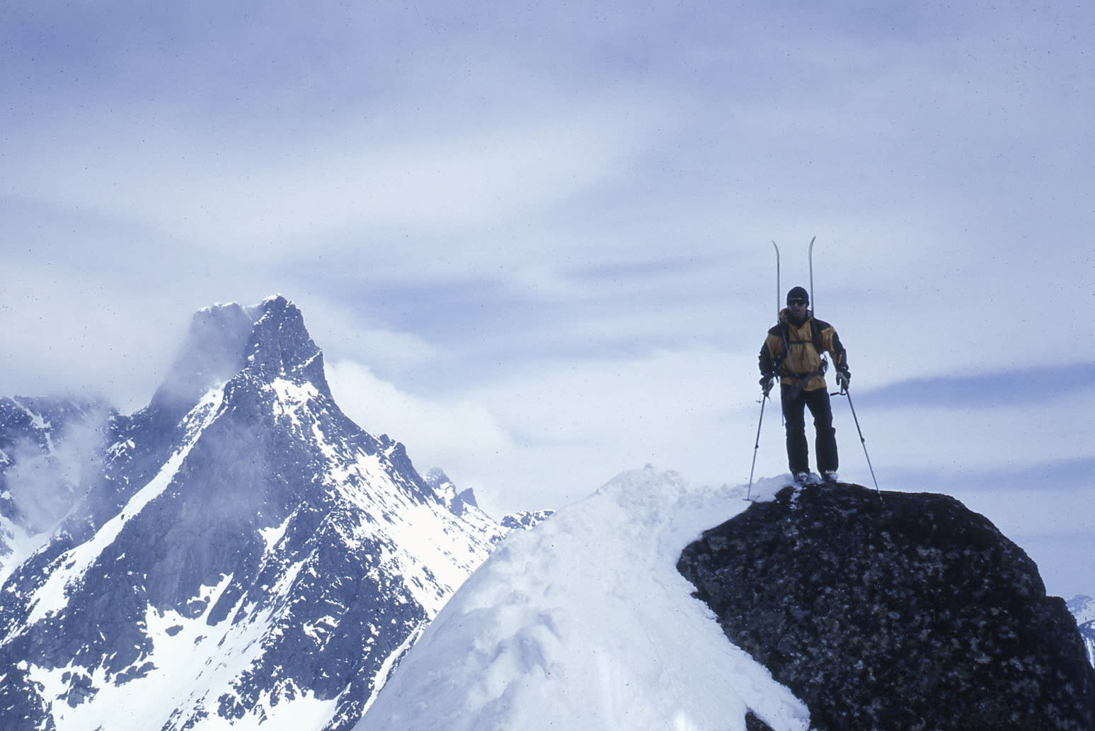 A person wearing Norrøna ski gear stands on a rocky outcropping with skis on their back and ski poles in hand. The snowy peak of a mountain is in the background.  The sky is mostly cloudy. 
