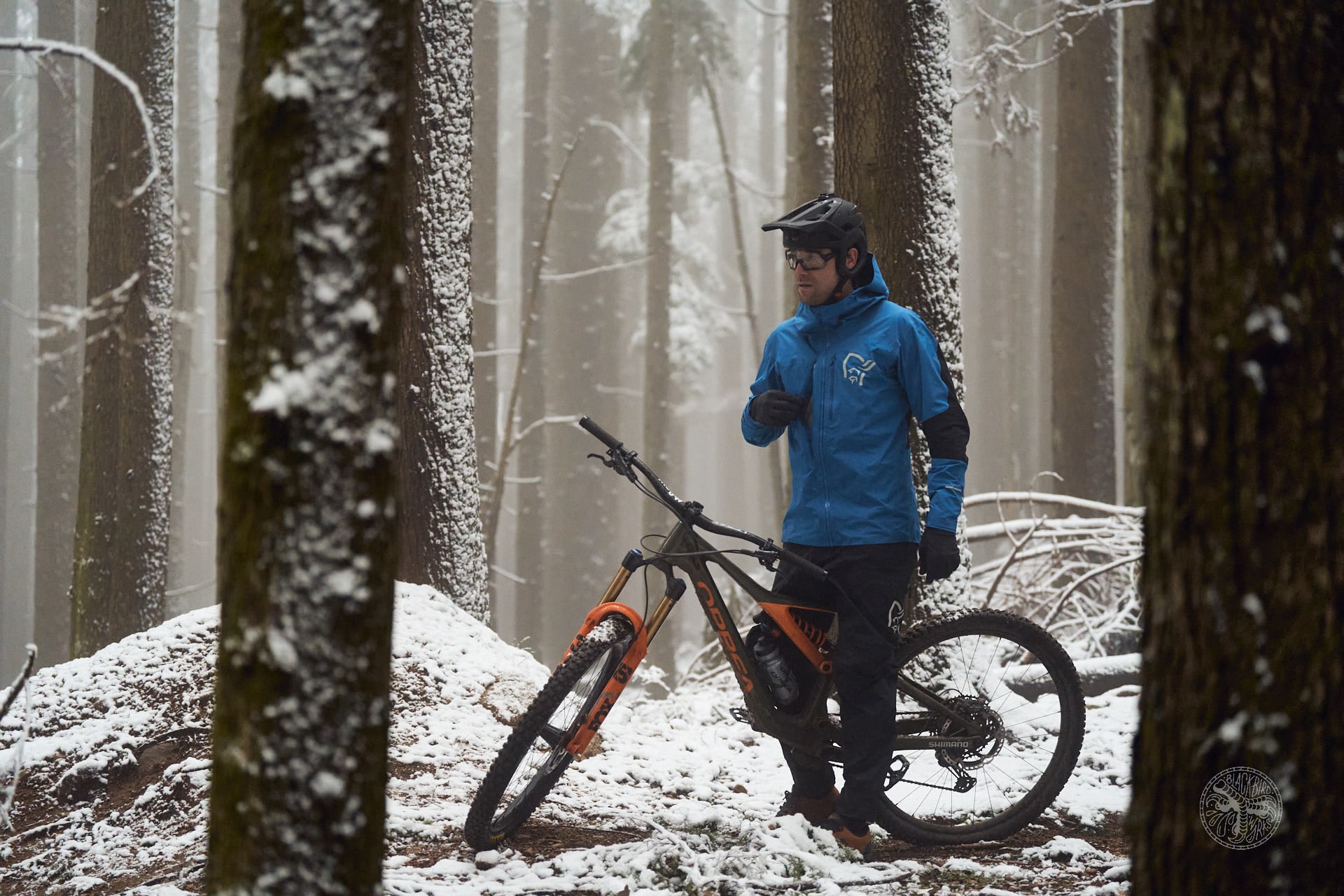 A man wearing a Norrøna jacket stands in a snowy forest, adjusting his jacket while leaning on a mountain bike. 
