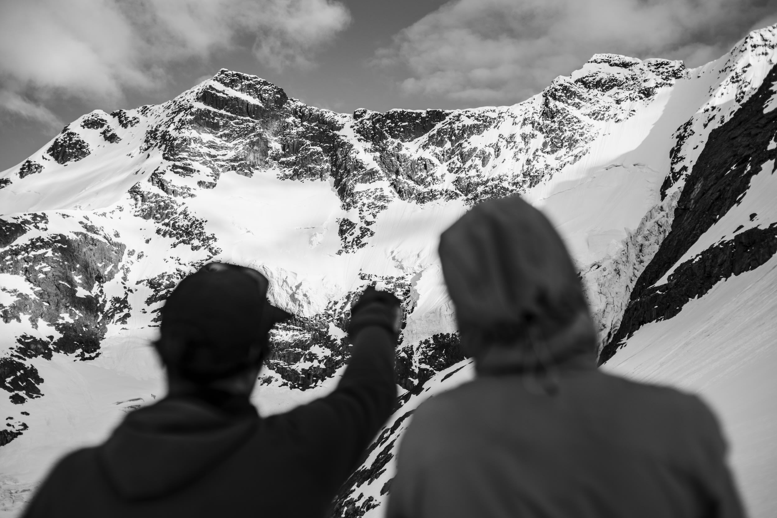 Two people, wearing Norrøna jackets, stand in front of a large snow-covered mountain range. They are silhouetted against the snow and the mountains. One of the people has a raised arm. The image is in black and white. 
