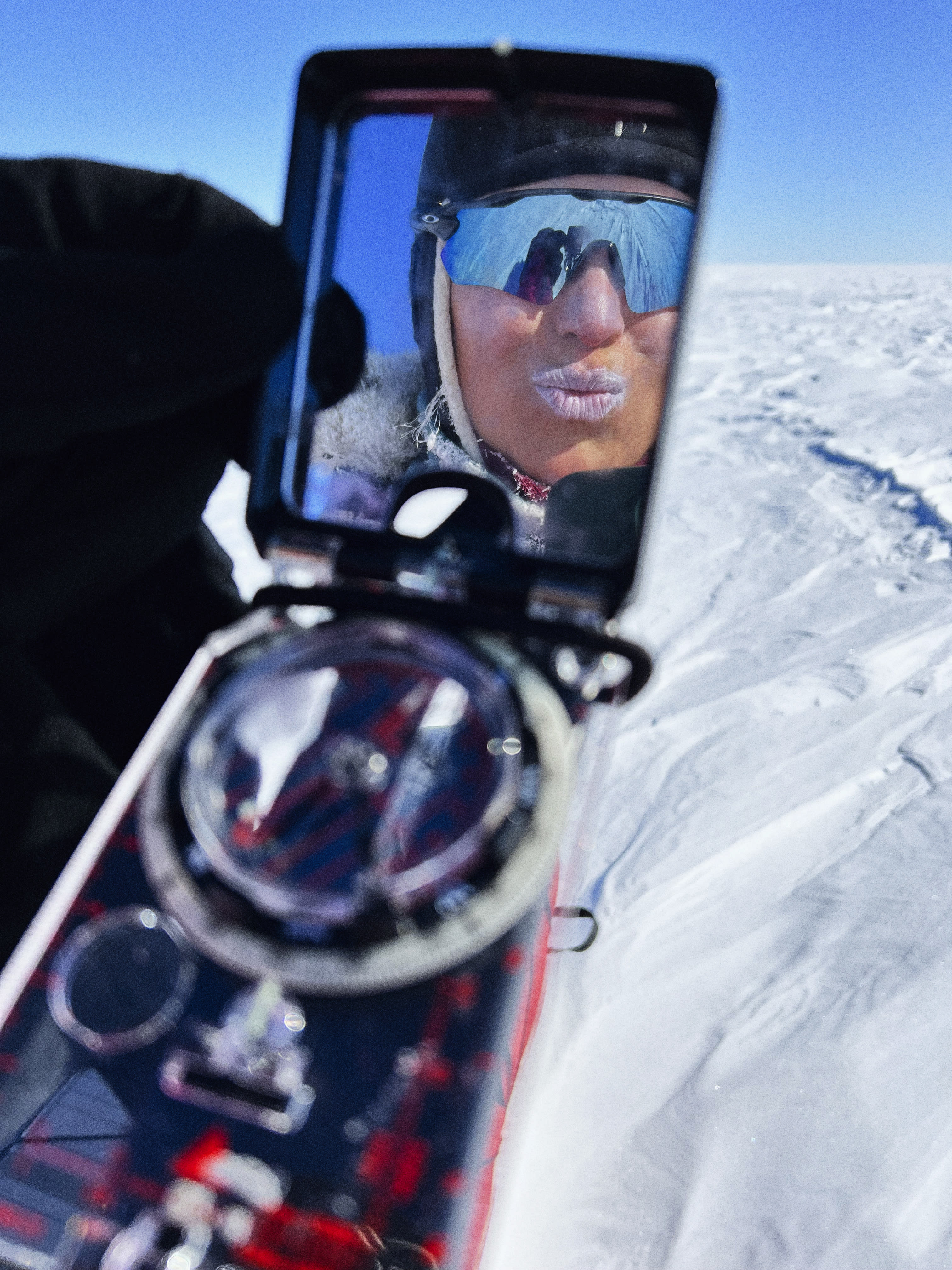 A person's reflection in a mirror, with a compass in the foreground. The person is wearing a black jacket and blue sunglasses. The background is a snowy landscape.