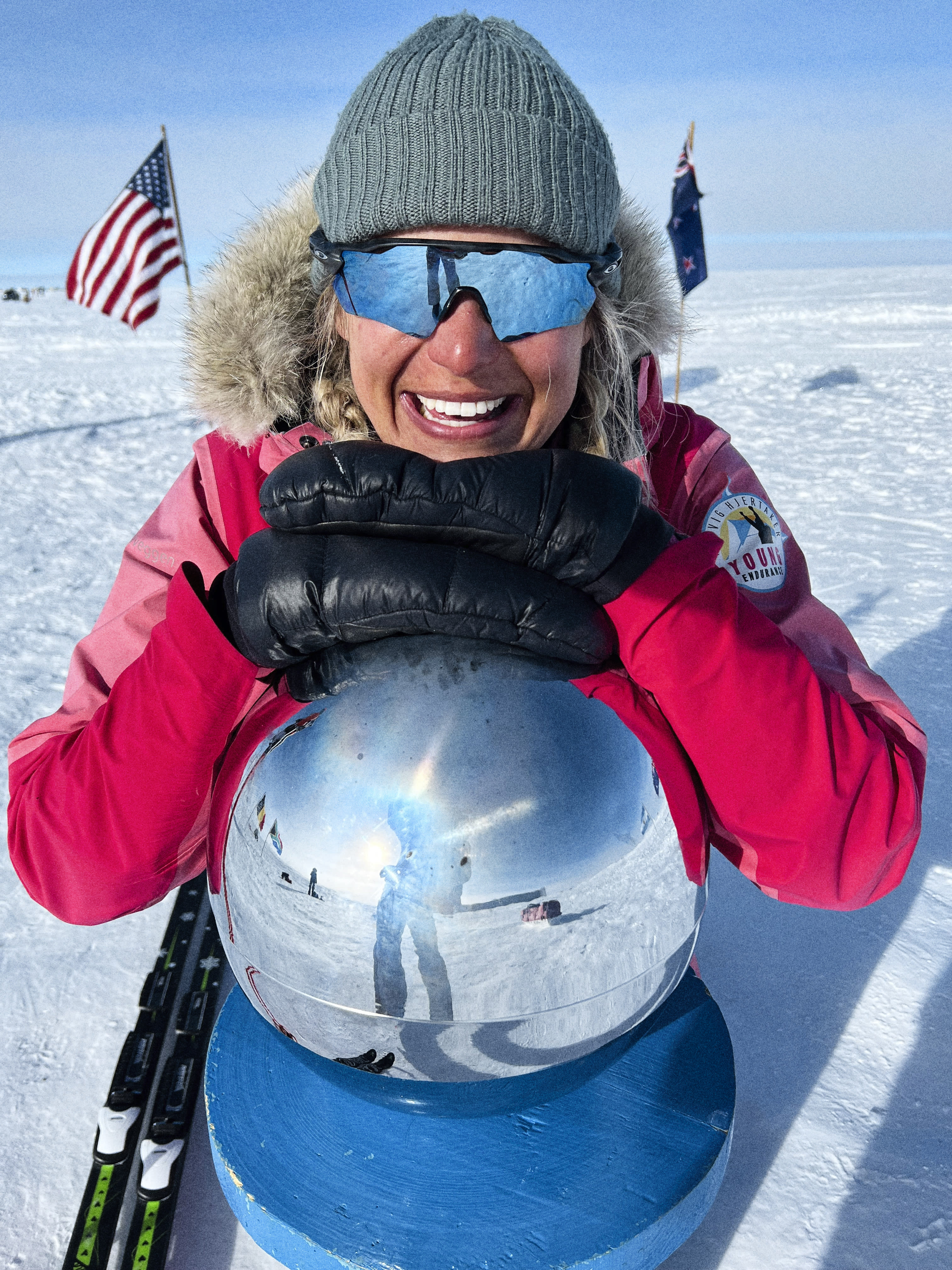 A woman in a pink jacket and grey beanie smiles while holding a large, reflective globe in front of her. The globe reflects the snow and sky behind her. A pair of skis lie at her feet. The image appears to be taken in Antarctica.